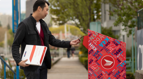 A man on a boardwalk opens the chute of a Canada Post street letterbox. He holds a flat rate box in his other hand.