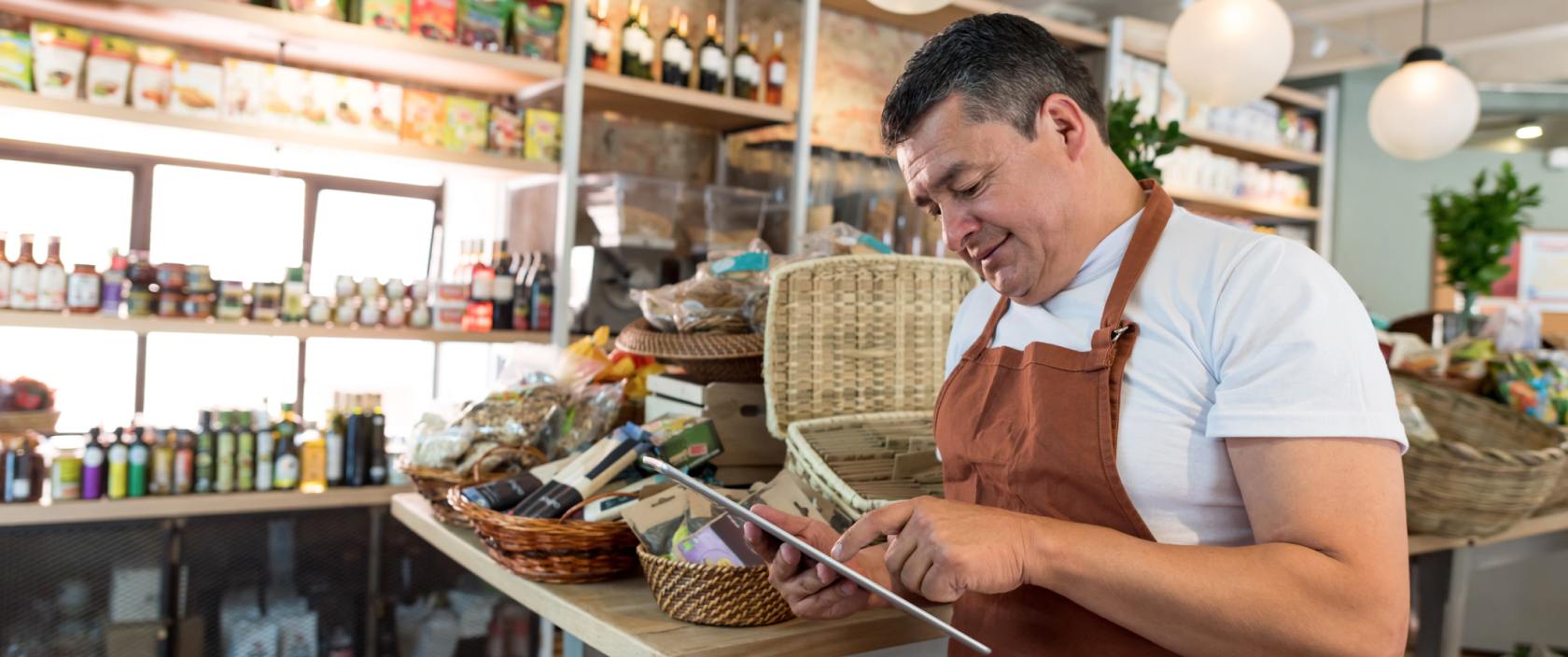 A man in an apron uses a tablet in a gourmet food shop.