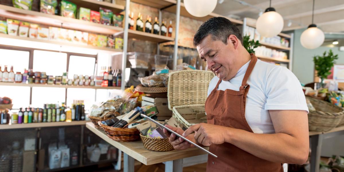 A man in an apron uses a tablet in a gourmet food shop.