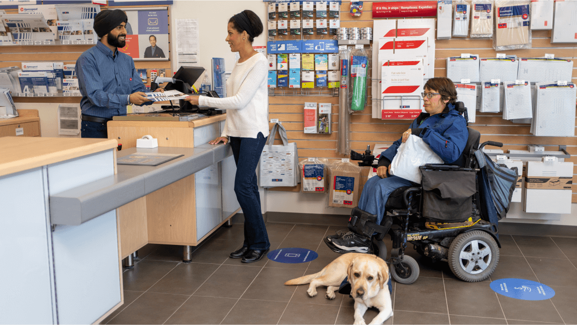 Une femme parle à un employé au bureau de poste. Une personne en fauteuil roulant avec un chien d’assistance attend en file.