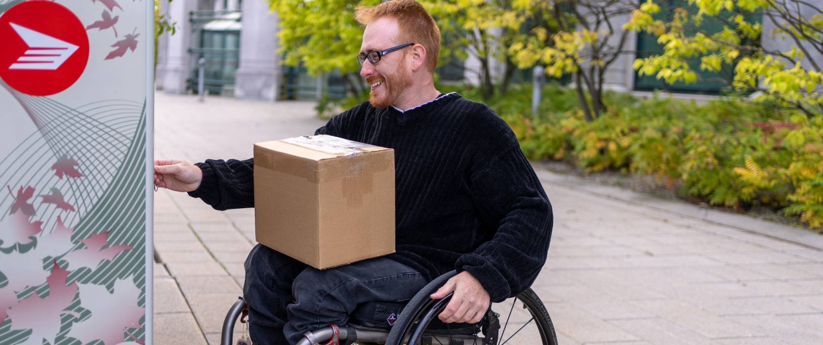 Un homme en fauteuil roulant ramasse un colis dans une boîte postale communautaire de Postes Canada.