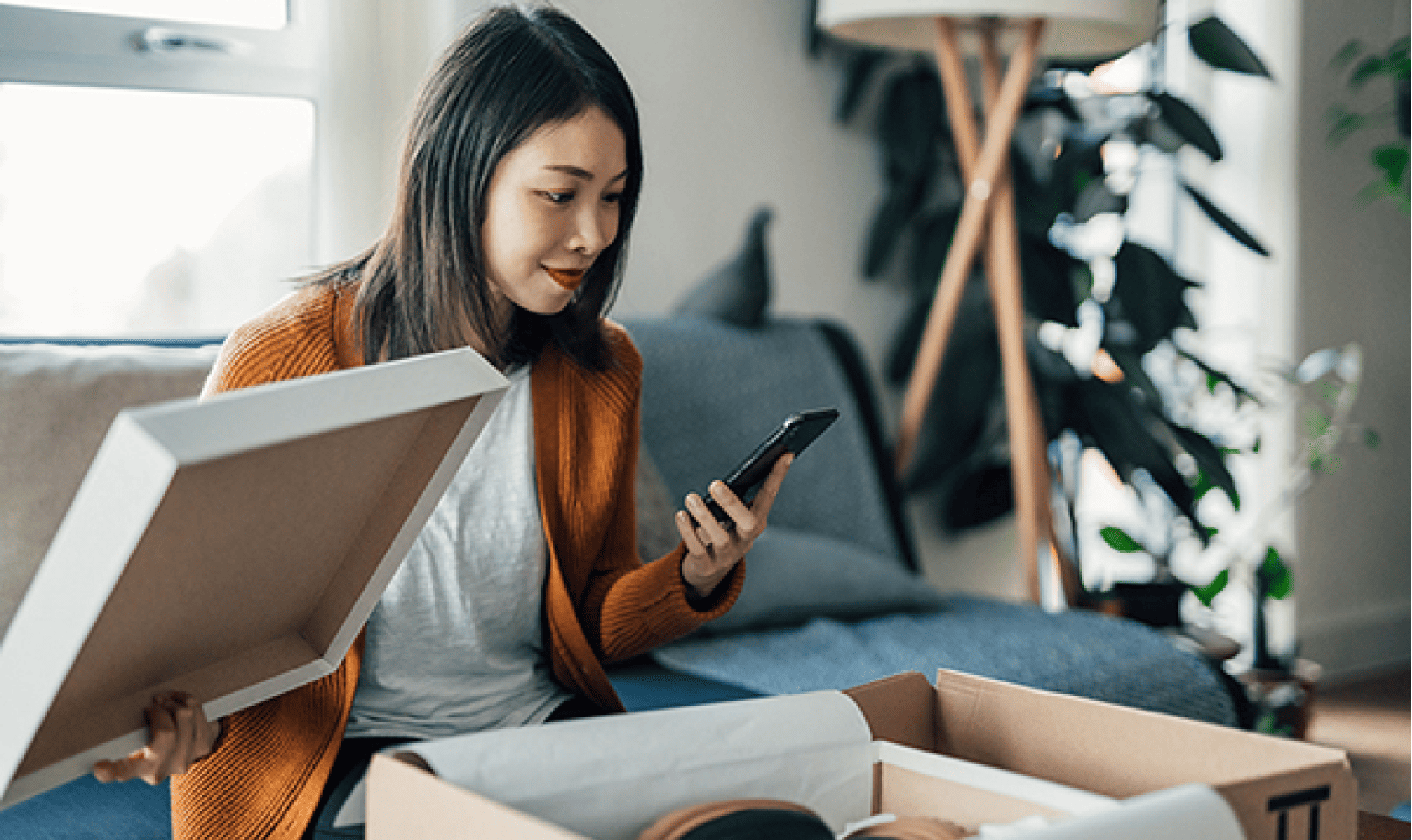 Dans son salon, une femme déballe un colis en consultant son téléphone.