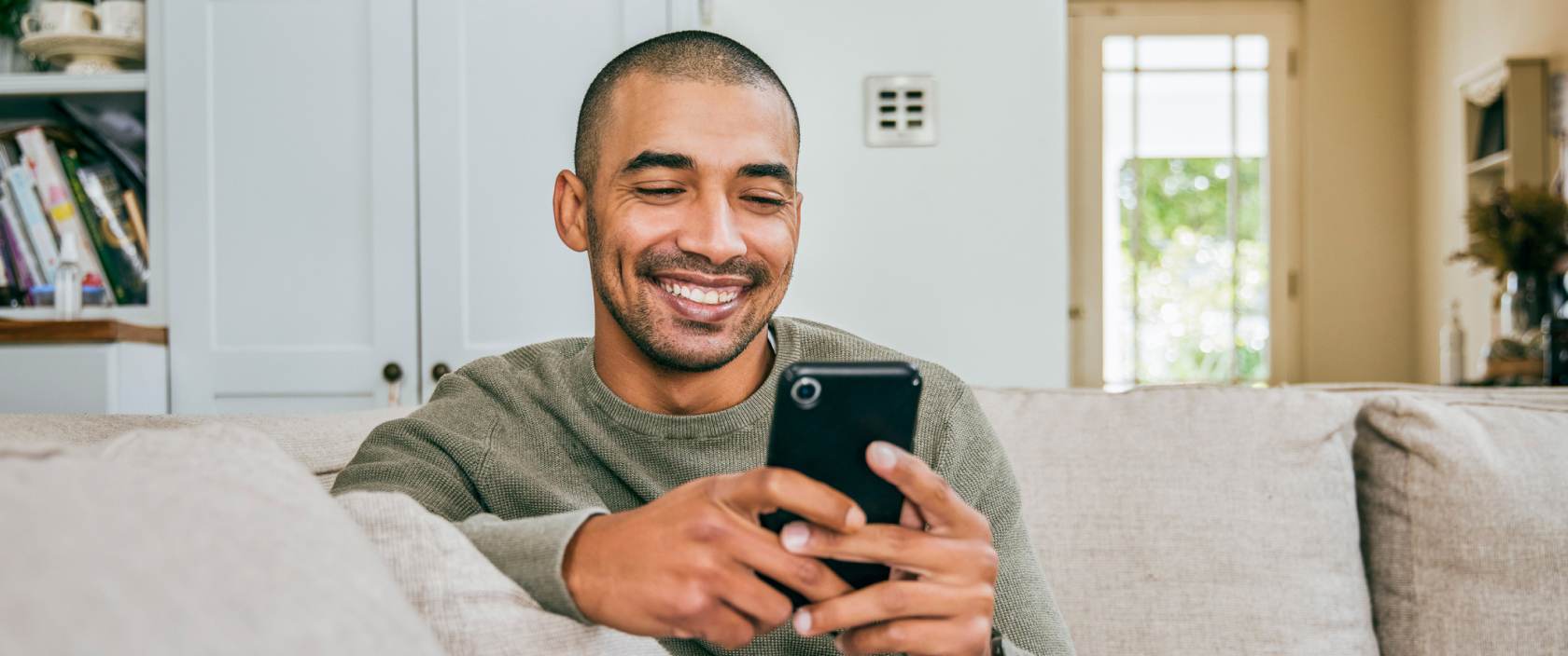 Un jeune homme sourit et fixe un téléphone cellulaire qui se trouve dans ses mains. Il est assis sur un canapé dans son salon.