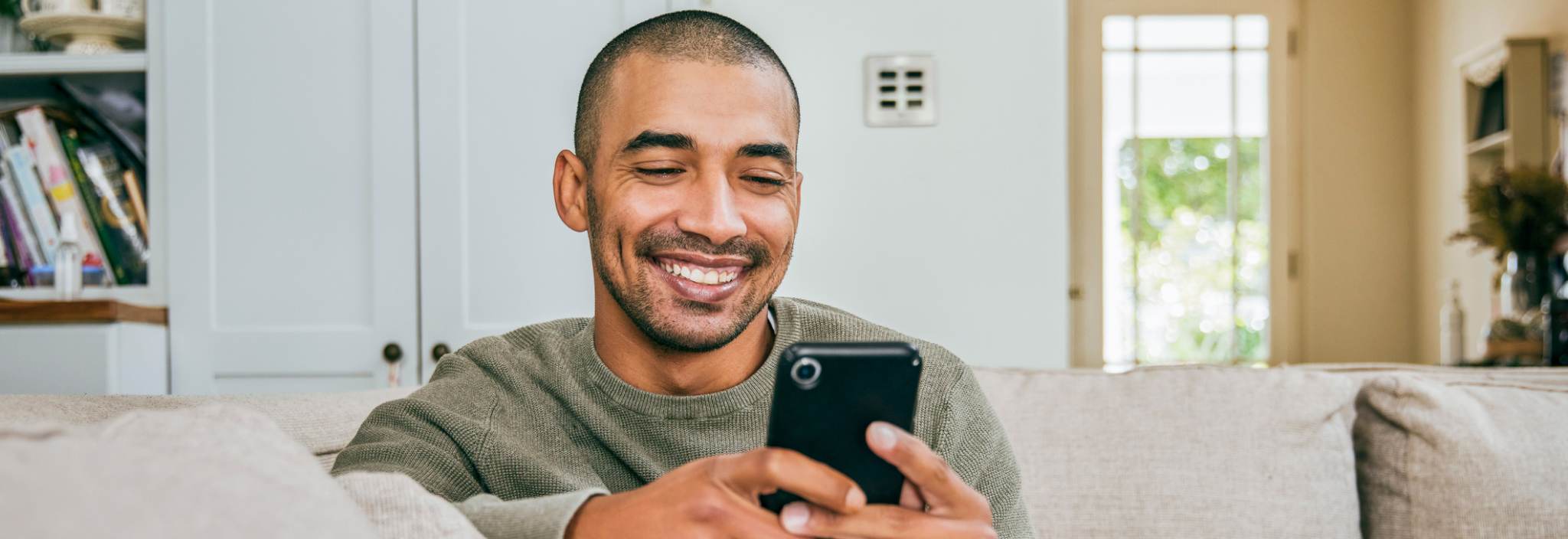 Un jeune homme sourit et fixe un téléphone cellulaire qui se trouve dans ses mains. Il est assis sur un canapé dans son salon.