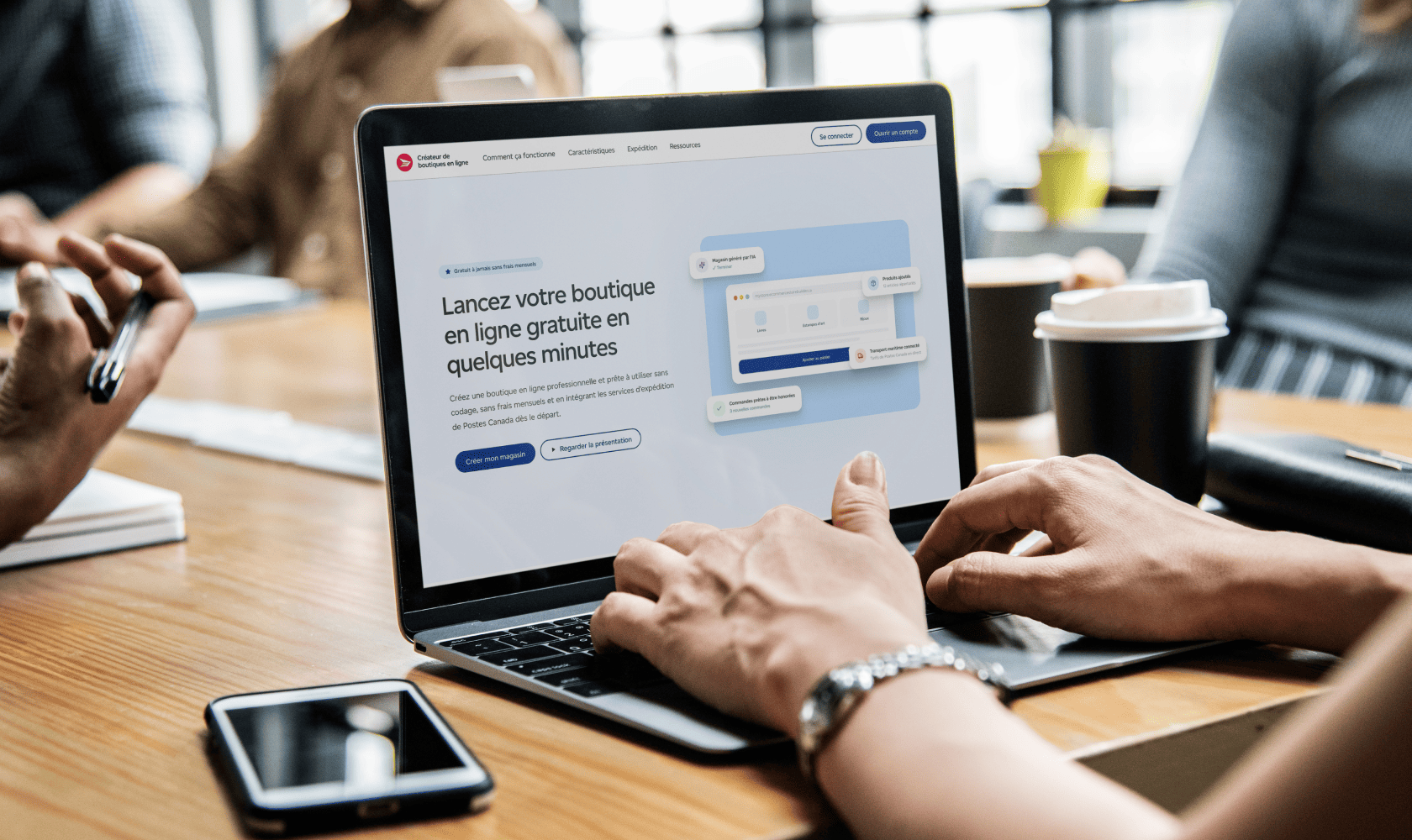Les mains d’une personne sur le clavier d’un ordinateur portable affichant la page de l’outil de création de boutiques en ligne. Sur la table de travail, il y a un téléphone intelligent, une tasse à café et des articles de bureau.