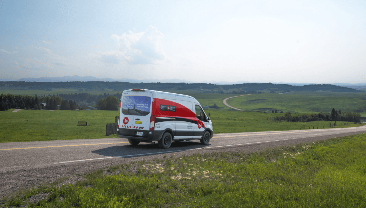 Une fourgonnette de livraison de Postes Canada roule sur une route rurale traversant des collines vertes sous un ciel gris.