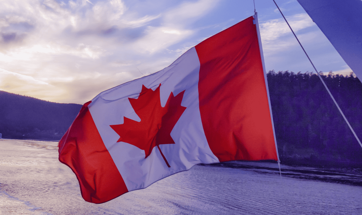 Un drapeau canadien flottant au vent, avec un plan d’eau et des collines boisées à l’arrière-plan.