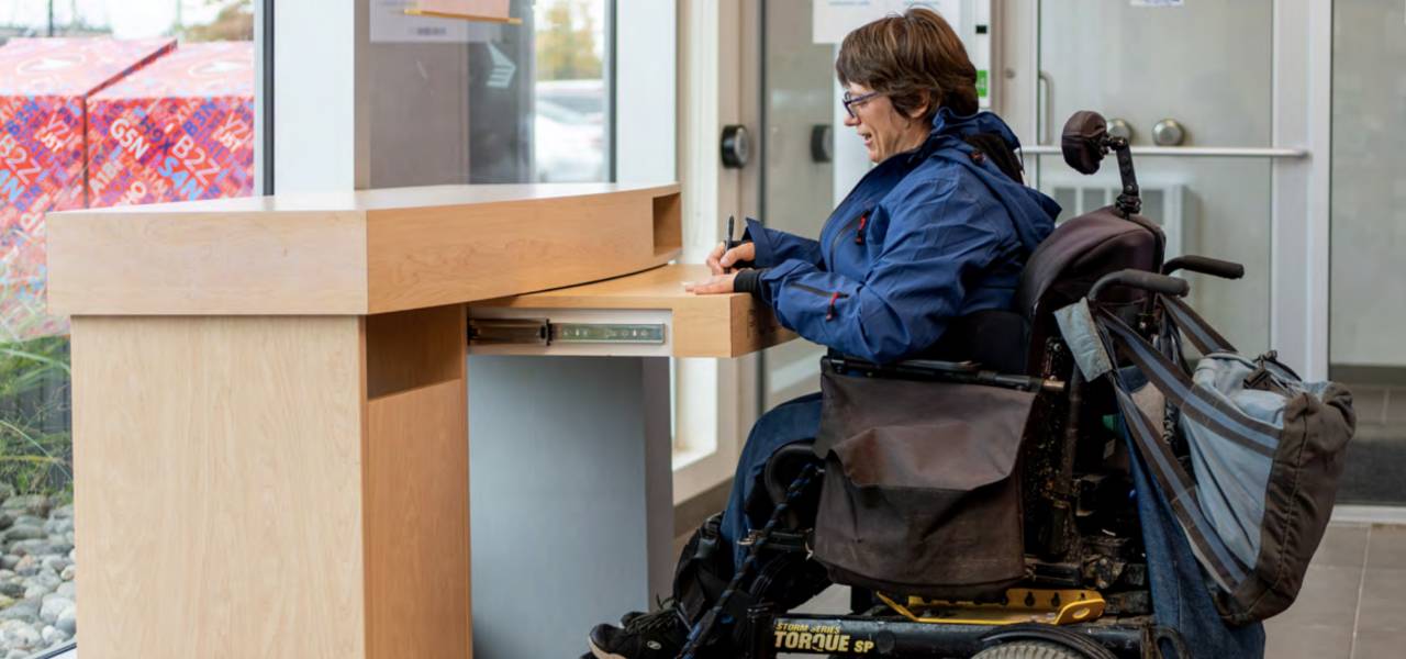 Une femme en fauteuil roulant remplit un formulaire à un bureau de poste accessible.