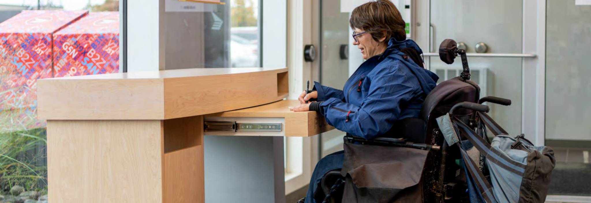 Une femme en fauteuil roulant remplit un formulaire à un bureau de poste accessible.