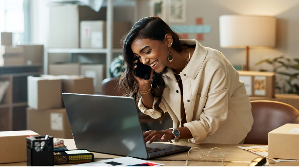 Une femme souriante parle au téléphone cellulaire et tape sur le clavier d’un portable près de boîtes d’expédition. Une femme souriante parle au téléphone cellulaire et tape sur le clavier d’un portable près de boîtes d’expédition.