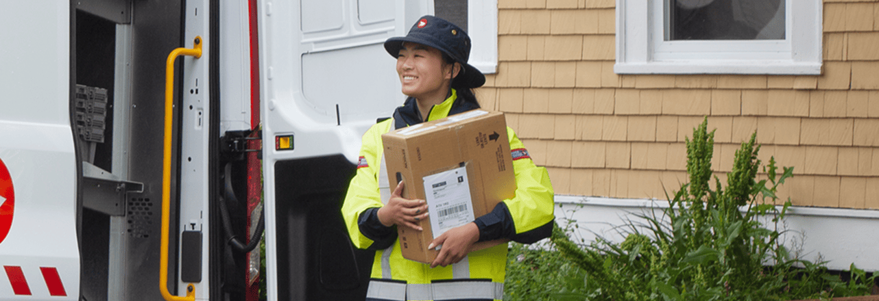 Un employé des postes en uniforme haute visibilité transporte un grand colis près d’un véhicule de livraison ouvert devant une maison.