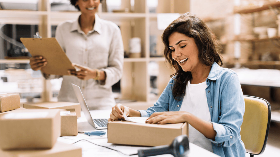 Une femme assise à un bureau appose une étiquette d’expédition sur une boîte. Une femme se tient près d’elle, une planchette à pince à la main.