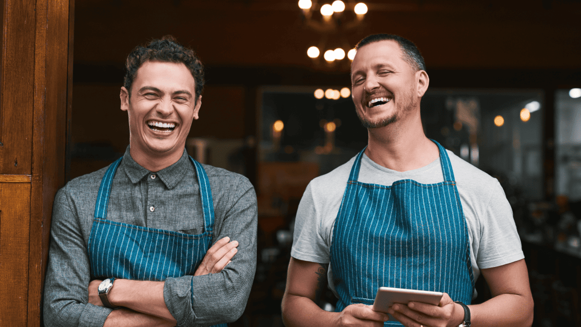 Deux hommes en tablier sourient et rient. L’un tient une tablette.