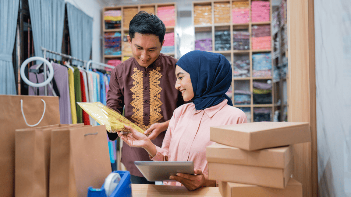 Une femme portant un couvre-tête est assise dans un magasin de vêtements. Un homme derrière elle lui tend une enveloppe.